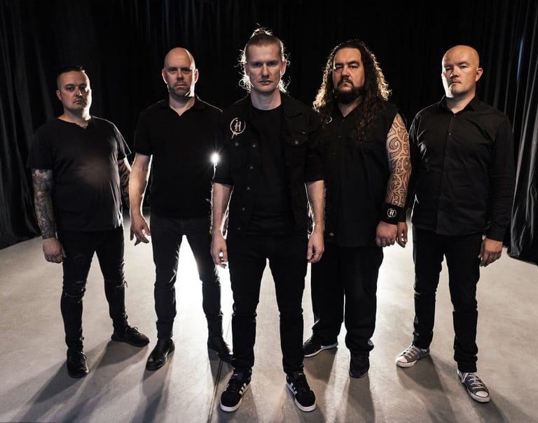 Five men in black clothing standing in a line against a dark background, professional band or group photo